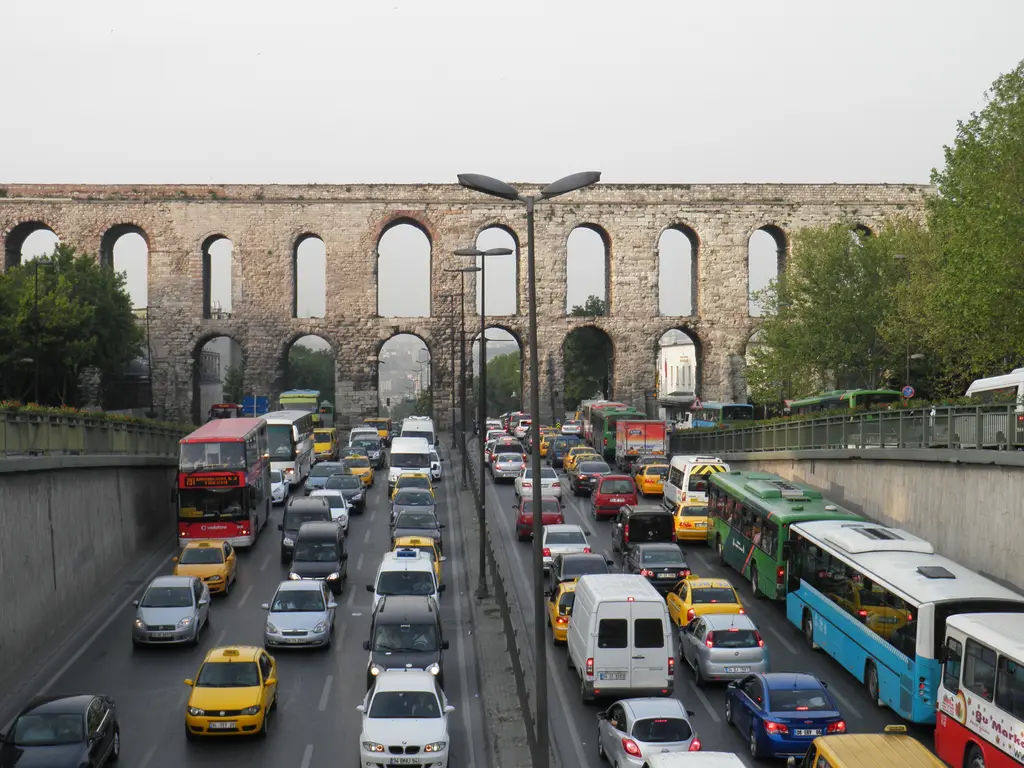 Bozdoğan Kemeri'nin altında yoğun trafik; çukur yolda kırmızı çift katlı otobüs, mavi beyaz belediye otobüsleri, onlarca sarı taksi ve arkada kemerin uzun arkadı.