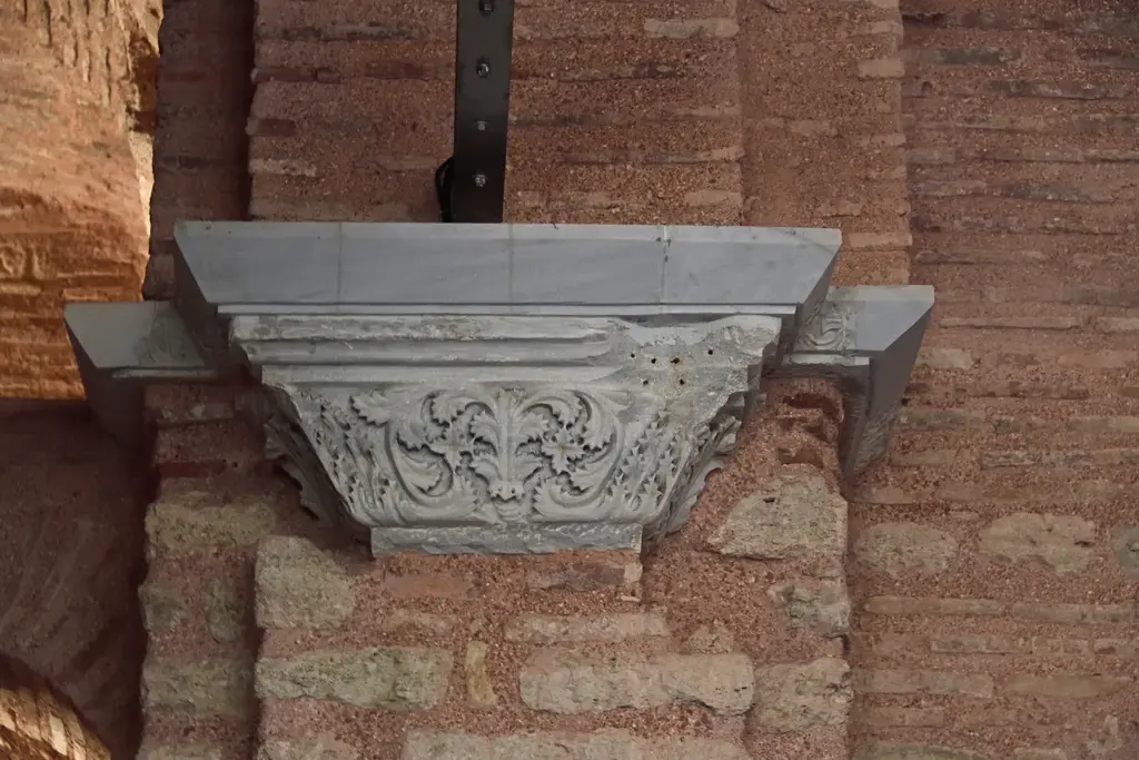 Fenari İsa Camii'nde tuğla duvara oturtulmuş beyaz mermer korniş bloğu; ön yüzünde kıvrımlı akant yaprağı kabartması, üstü düz plaka, üstünde metal bağlantı.