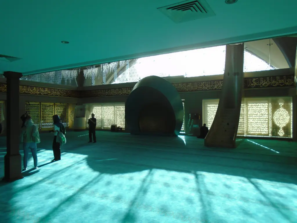 Şakirin Camii'nde harim; turkuaz halı üzerinde dolaşan ziyaretçiler, mihrap, minber ve geniş cam pencereler.