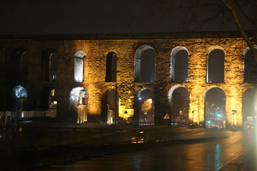 Bozdoğan Kemeri yağmur sonrası gece; sarı ışıkla aydınlanmış gövde puslu, gözlerden geçen ışık huzmeleri sise vuruyor, ıslak asfalt lambaları ve altın yansımaları aynalıyor.