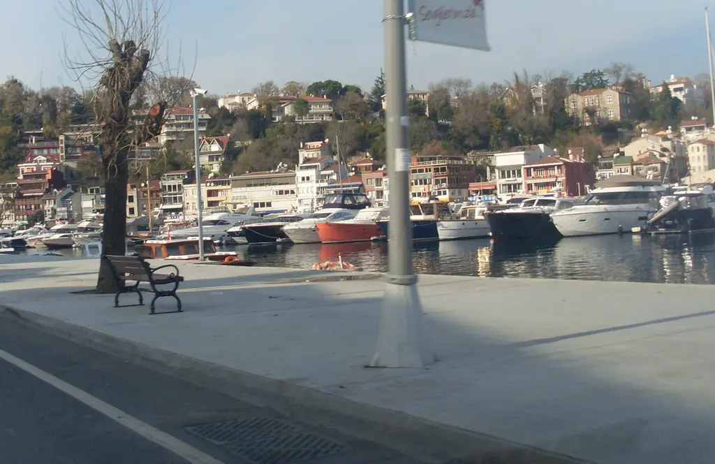 Tarabya'da marina önündeki geniş kaldırım; bank, ağaç, sokak lambası ve arkada tekneler ile yamaçtaki evler.