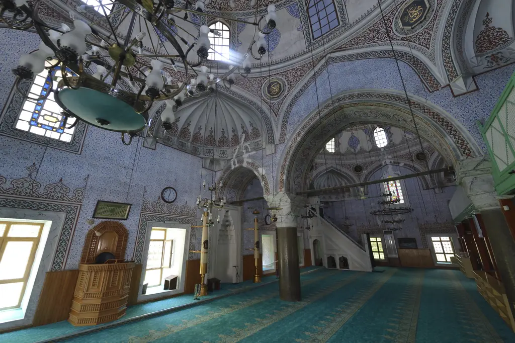 Haseki Sultan Camii'nin harimi; yeşil halı, taş sütunlar üstünde kemerler, ahşap kürsü, pencereler ve avizeler.