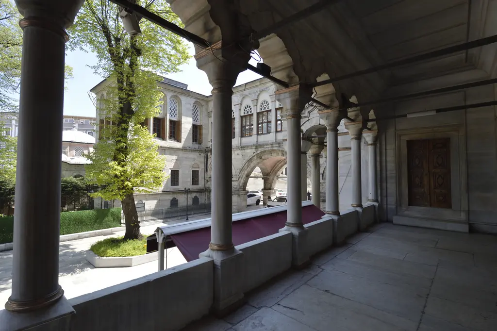 Nuruosmaniye Camii'nde hünkâr kasrına giden üstü örtülü revak; sıra sütunlar, mor tente, dışarıda yeşillenmiş ağaç ve kasrın büyük pencereli cephesi.