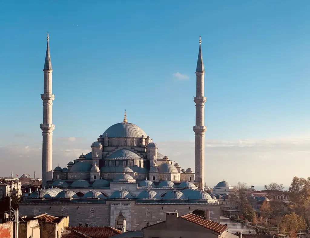 Fatih'te Fatih Camii'nin çatılardan görünümü; gri kurşun ana kubbe, kademeli inen yarım kubbeler, iki yanda ince beyaz minareler ve önde kırmızı kiremitli evler.
