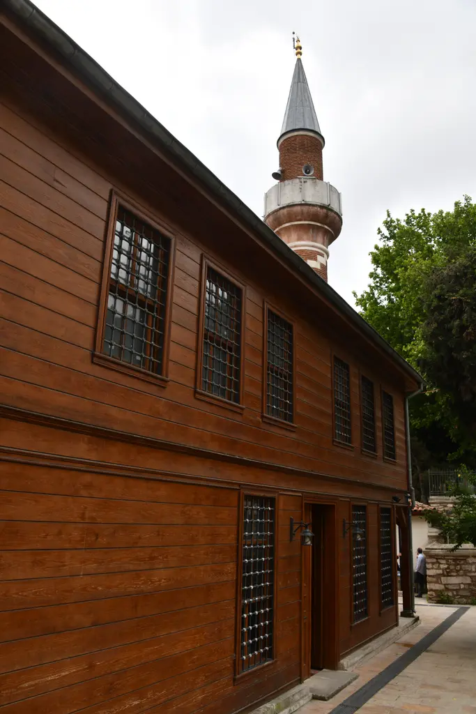 Şah Sultan Camii'nde ahşap bina; iki katlı kahverengi cephe, demir şebekeli pencereler, minare ve bulutlu gökyüzü.