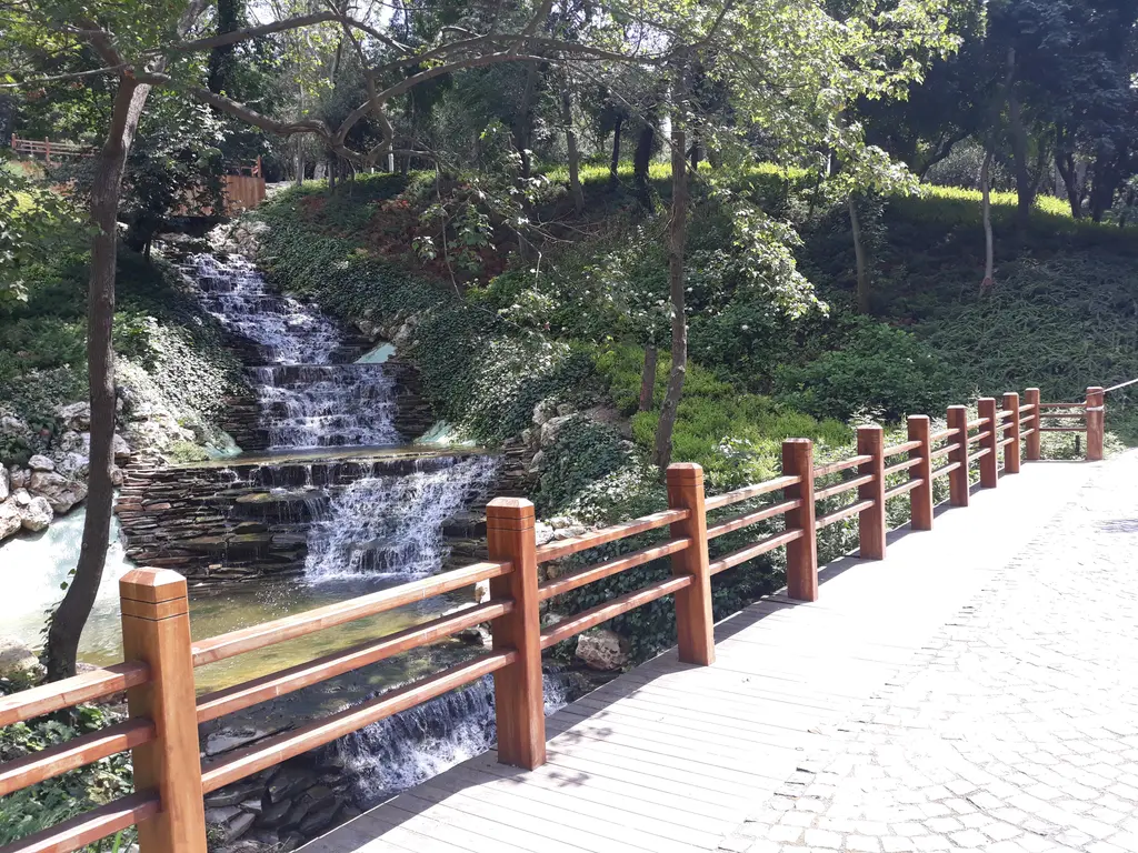 Yıldız Parkı'nda bir şelale; taşların üstünden akan su, sık yeşillik ve önünde kırmızı ahşap korkuluklu bir yol.
