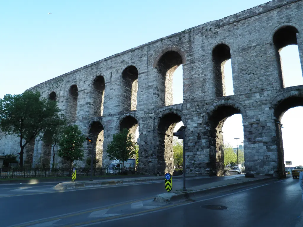 Bozdoğan Kemeri alacakaranlıkta perspektifle uzaklaşıyor; yakın uçta iki dev kemer gözü, açık gri taş, dipte yeşil ağaç sırası, boş ıslak asfalt yol ve turkuaz gökyüzü.