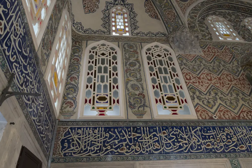 Sultan Ahmed Türbesi'nde duvar ve pencereler; alçı şebekeli renkli camlar, kalem işli yüzeyler ve altta lacivert çini yazı kuşağı.