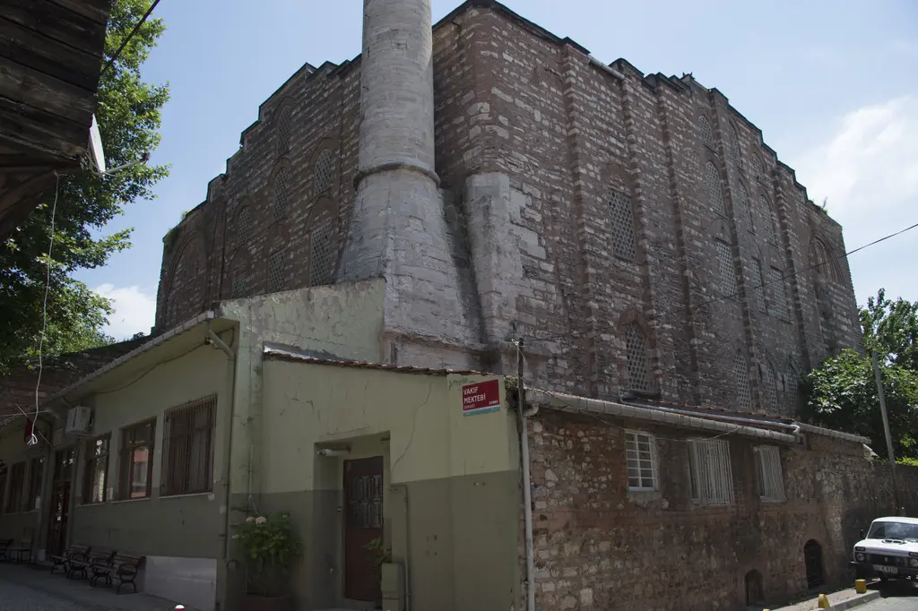 Gül Camii'nin batı köşesi; yüksek tuğla gövde, beyaz minare, önde yeşil boyalı tek katlı yapı ve sokak.