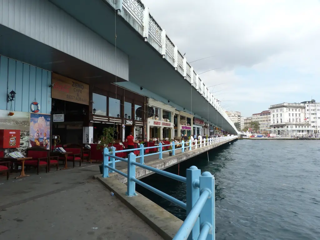 Galata Köprüsü'nün alt katında boş masalar; mavi korkuluk, köprü tabliyesinin gölgesi, Haliç ve karşı kıyıdaki binalar.