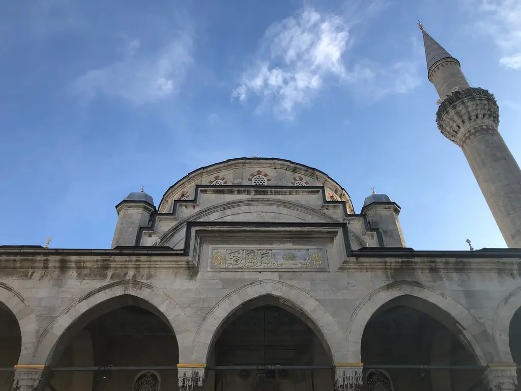 Şişli Camii'nin giriş cephesine aşağıdan bakış; kemerli revak, pencereli kubbe kasnağı, köşe kuleleri ve sağda yükselen minare.