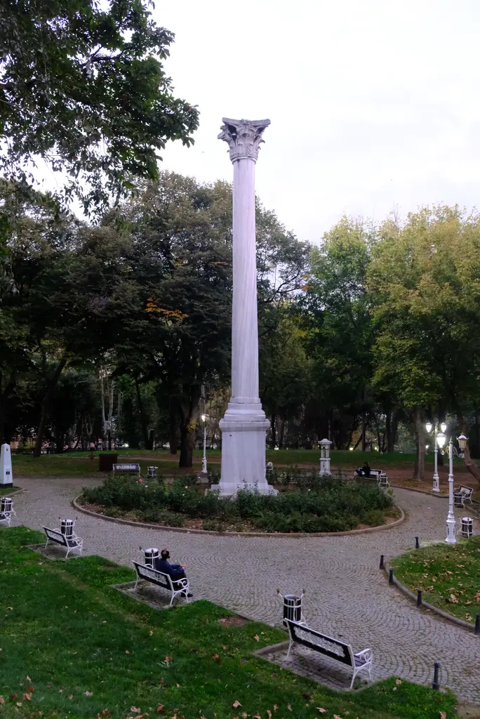Gotlar Sütunu ve çevresindeki çiçek tarhı; parke taş halka yol, banklar, lamba direkleri ve arkada sık yeşil ağaçlar.
