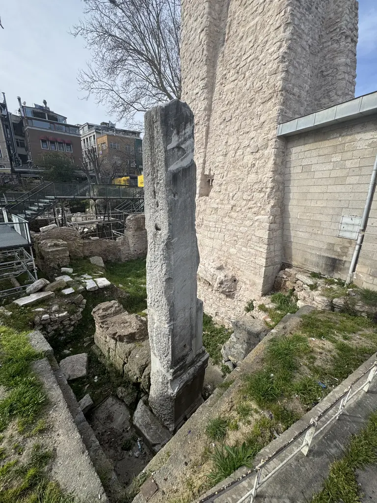 Milyon Taşı ve su terazisinin açık renk taş cephesi; mermer ayak, dibinde otlar, solda kazı alanı ve arkada eski binalar.