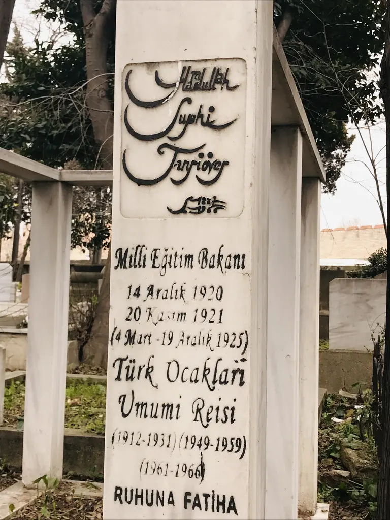 Uzun beyaz mezar taşı; üstünde ay yıldız kabartması, gövdesinde ad ve görevlerin yazılı olduğu uzun kitabe, arkada selviler.