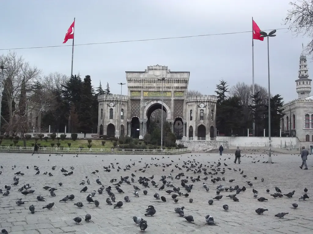 Kış gününde Beyazıt Meydanı ve kule; çıplak ağaçlar, taş zeminde güvercinler, üniversite kapısı ve arkasında kulenin silueti.