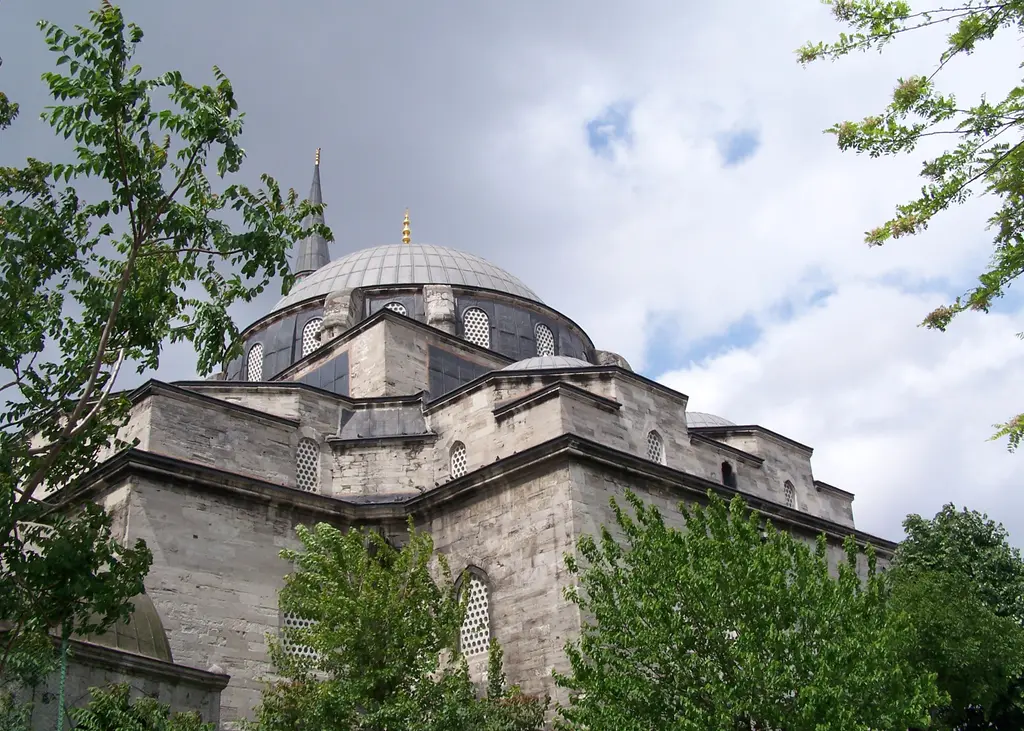 Gazi Atik Ali Paşa Camii; yeşil ağaçlar arasından yükselen kesme taş gövde, kademeli kubbeler, kurşun ana kubbe ve bulutlu gökyüzü.