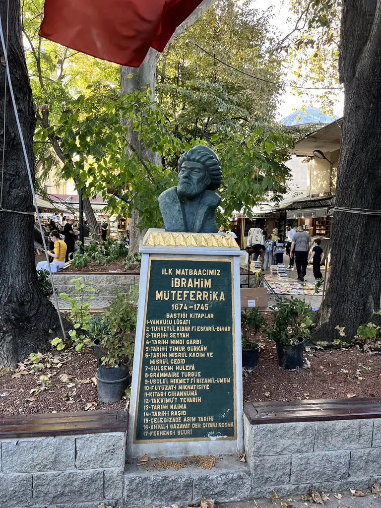 Sahaflar Çarşısı'nda İbrahim Müteferrika büstü; yeşil patinalı bronz baş, beyaz mermer kaide, kaidede yaldızlı kitabe, arkada çınar gövdeleri ile bir caminin kurşun kubbesi.