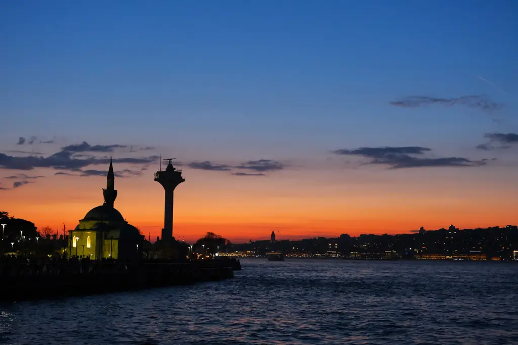 Gün batımında Şemsi Paşa Camii; turuncu gökyüzü önünde kubbe ve minare silueti, yanda bir kule ve deniz.