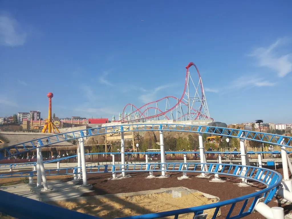 Vialand'de mavi beyaz raylı alçak bir ünite ve arkada yükselen kırmızı roller coaster; solda kırmızı toplu yüksek bir direk.