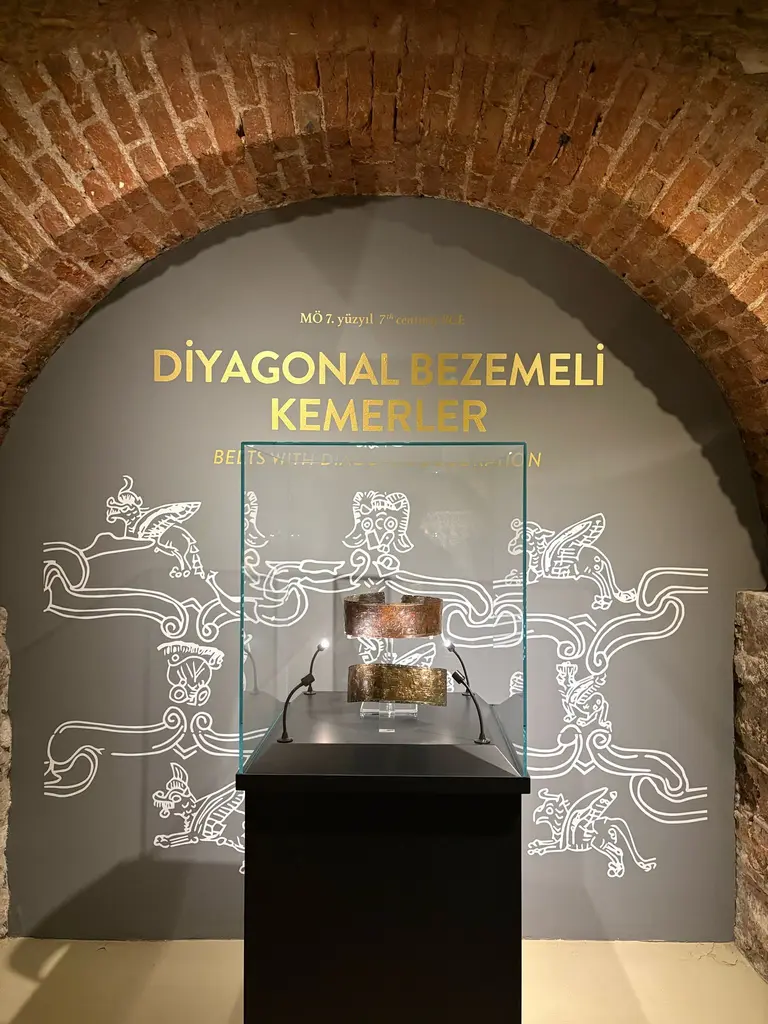 Tuğla kemerli bir nişte Diyagonal Bezemeli Kemerler sergisi; gri duvarda altın harfli başlık, beyaz çizgili hayvan figürleri ve cam vitrin.