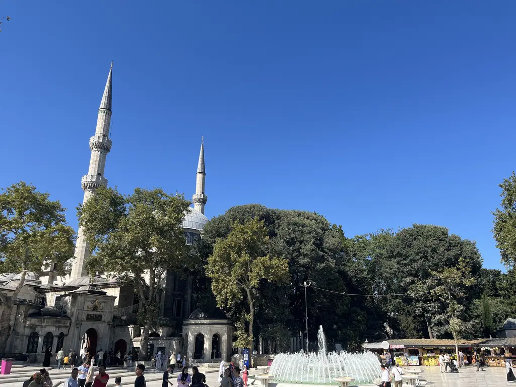 Eyüpsultan meydanı; mavi gökyüzüne yükselen iki minare, ağaçlar, fıskiyeli havuz ve dolaşan ziyaretçiler.