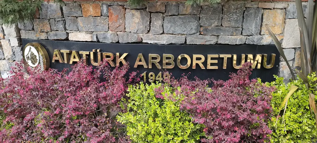 Atatürk Arboretumu'nun girişi; taş duvarda yaldızlı harflerle yazılmış ad ve önünde mor yapraklı çalılar.