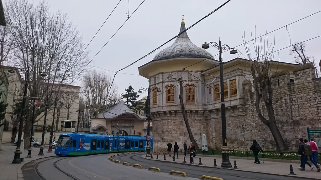 Bulutlu bir günde Alay Köşkü ve Alemdar Caddesi; soğan kubbeli köşk, sur, Gülhane kapısı, virajdaki mavi tramvay ve yürüyen insanlar.