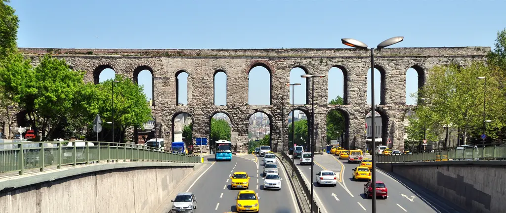 Bozdoğan Kemeri geniş panoramada; iki katlı kemer dizisi baştan sona uzanıyor, altta çukur bulvarda sarı taksiler ile mavi otobüs, solda cam korkuluklu üstgeçit.