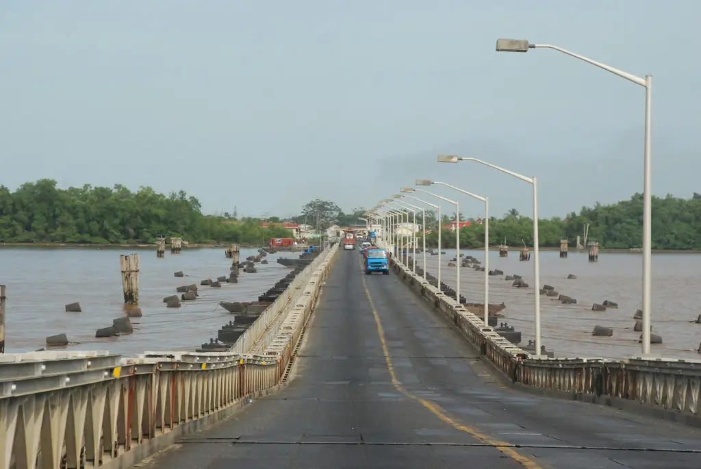 Guyana'da geniş, çamurlu kahverengi bir nehri aşan uzun yüzer köprü; üzerinden geçen kamyonlar, sıra sıra sokak lambaları ve iki yakadaki alçak yerleşim.
