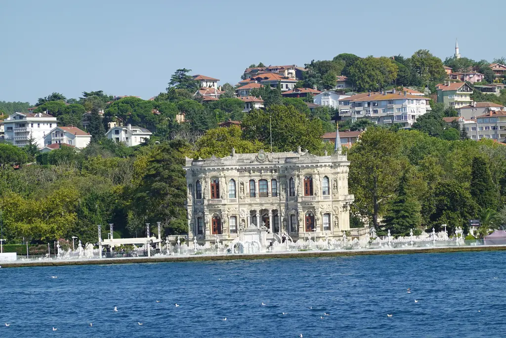 Denizden Küçüksu Kasrı; Boğaz kıyısında beyaz kasır, önünde parmaklıklı rıhtım, arkada ağaçlar ve yamaçtaki evler.
