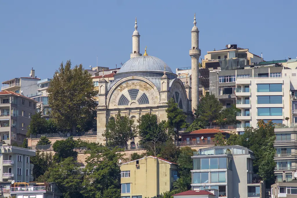 Cihangir Camii'nin yamaçtaki görünümü; gri kubbe, iki minare, ağaçlar ve çevresinde renkli cepheli apartmanlar.