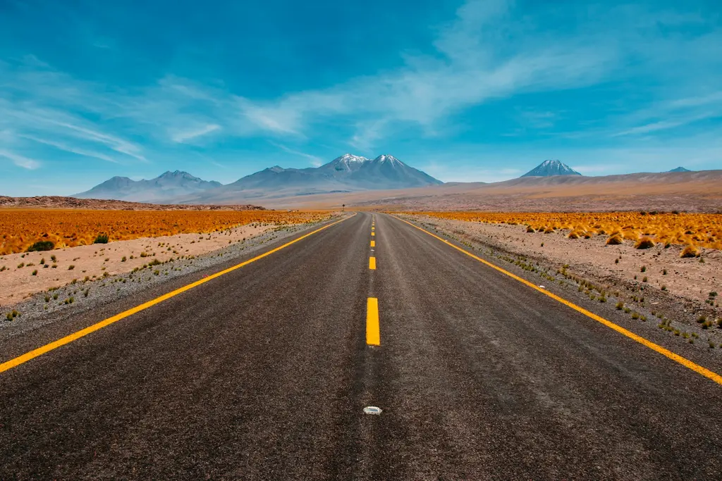 Şili'de Atacama Çölü'nde ufka doğru uzanan sarı çizgili asfalt yol; iki yanda kuru bozkır ve uzakta karlı volkan zirveleri, parlak mavi gökyüzü.