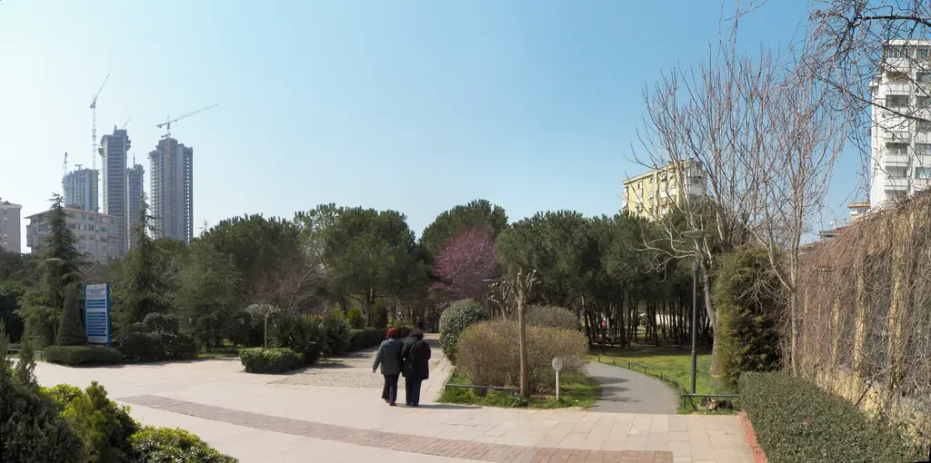 Göztepe'de Özgürlük Parkı; geniş taş yürüyüş yolu, çam ağaçları, çiçekli erguvan ve arkada gökdelenler.