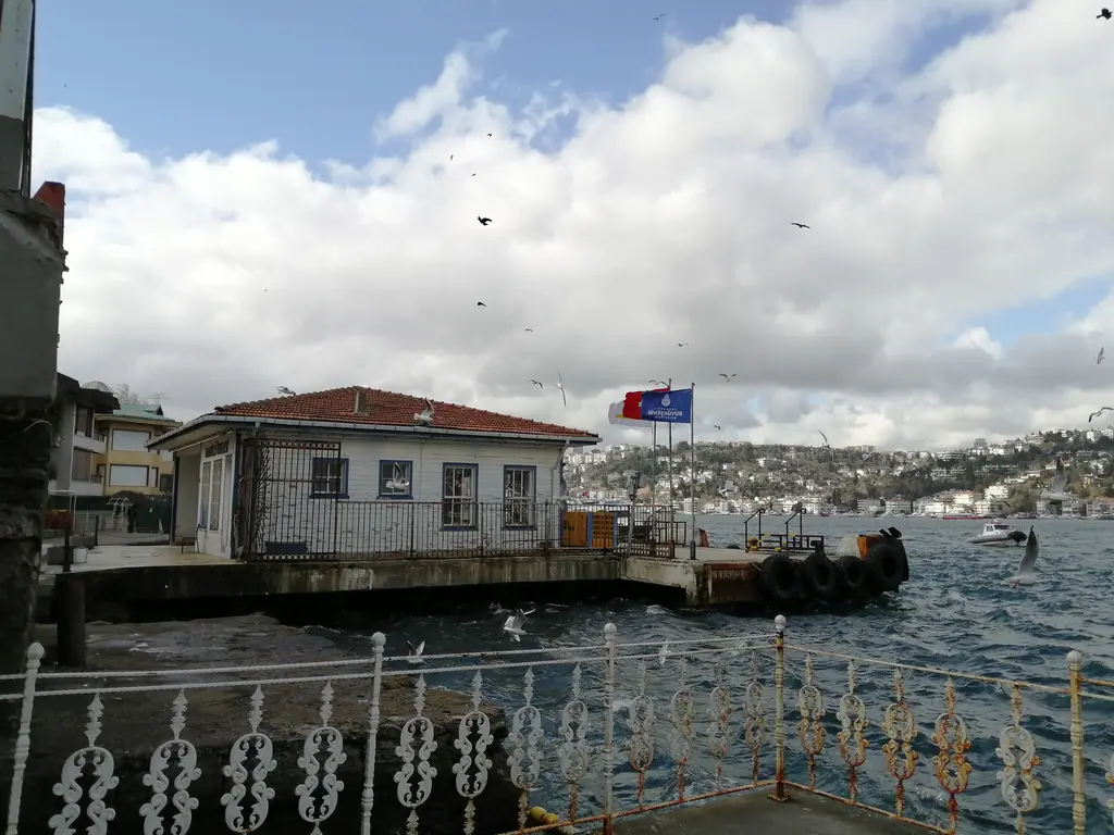 Kandilli İskelesi; deniz kenarında ahşap iskele yapısı, önünde bayrak, arkada Boğaz ve karşı kıyı.