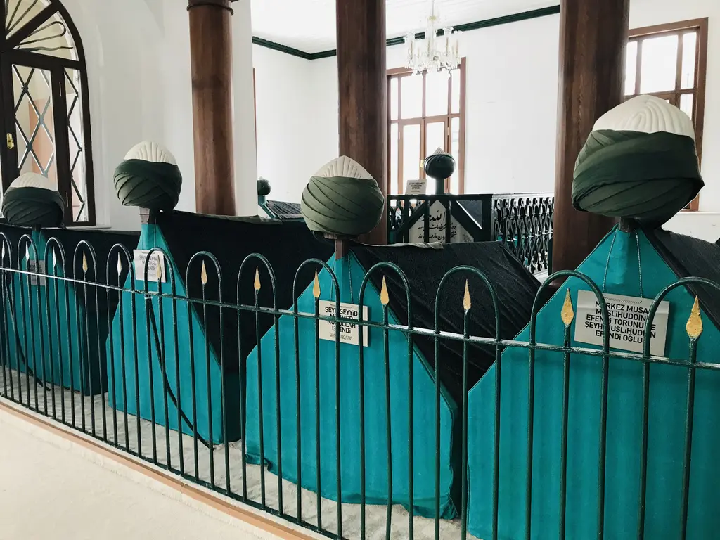 Türbe içinde turkuaz parmaklıkların ardında sıralanan sandukalar; yeşil örtüler, sarıklı başlıklar ve tavandan sarkan zincirler.