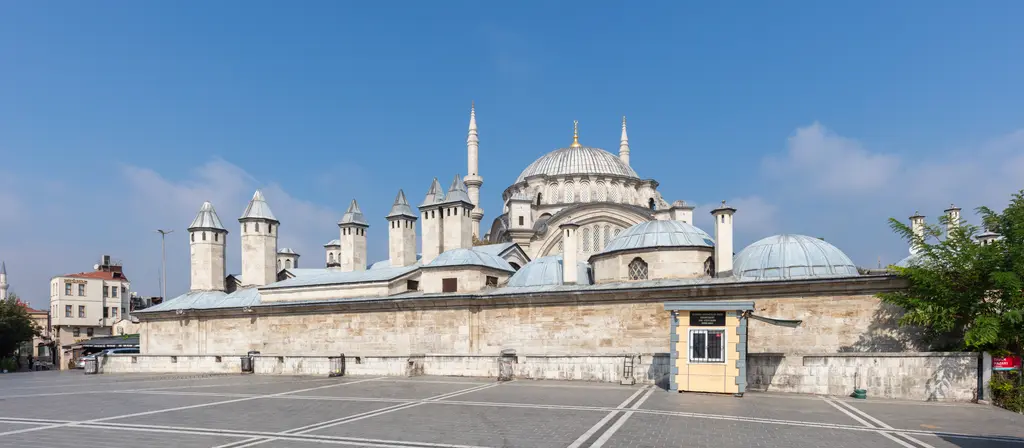 Nuruosmaniye Camii'nin güney tarafı; önde geniş boş taş meydan ile avlu duvarı, arkada kubbe, iki minare ve külliye yapılarının külahları, sert öğle ışığı.