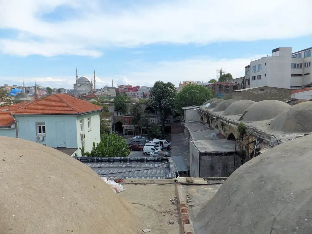 Büyük Valide Han'ın çatısından bakış; önde iki büyük sıvalı kubbe ve sağda kubbe sırası, aşağıda dükkânlı sokak, uzakta kubbeli ve iki minareli bir cami.