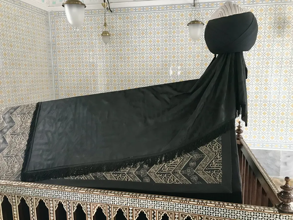 Türbenin içinde koyu yeşil örtülü sanduka; baş tarafında sarıklı ahşap başlık, arkada çini kaplı duvar ve zeminde renkli mozaik.