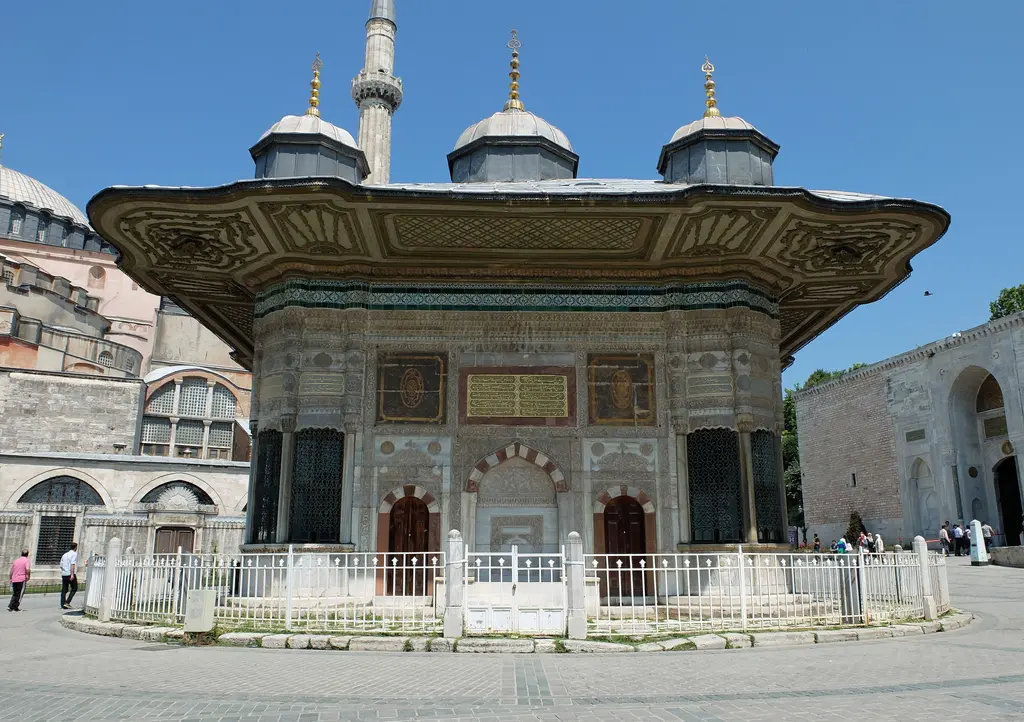 Çeşme Ayasofya yönünden; sağda Ayasofya'nın duvarları ve minaresi, ortada çeşmenin saçaklı gövdesi, önde taş döşeli meydan.