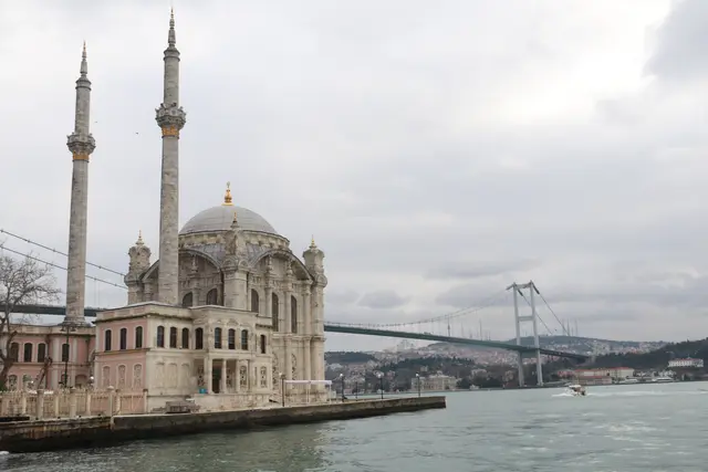Beşiktaş'ta Ortaköy Camii; kıyıdaki kubbeli beyaz cami, iki ince minare, arkada Boğaz'ı aşan asma köprü ve bulutlu gökyüzü.