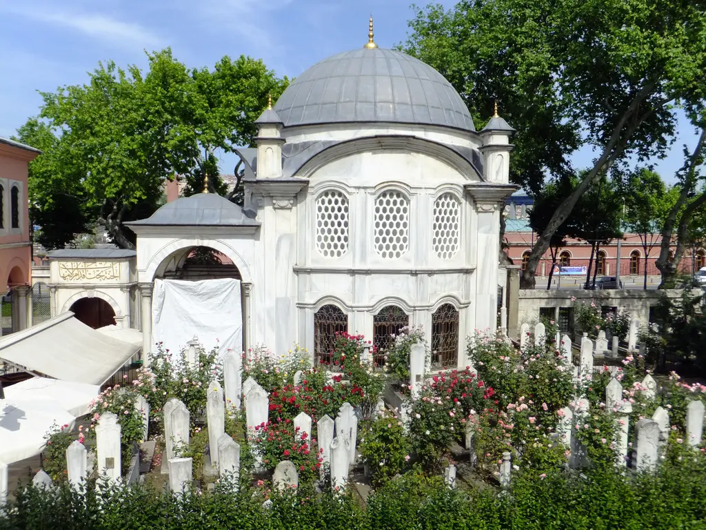 Şah Sultan Türbesi güneyindeki kabristandan; sık dizili beyaz mezar taşları, kırmızı güller, revak ve kubbeli türbe.