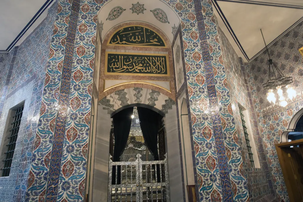 Eyüp Sultan Türbesi'nin içi; çini kaplı duvarlar, hat levhaları, kemerli pencereler ve ziyaretçiler.