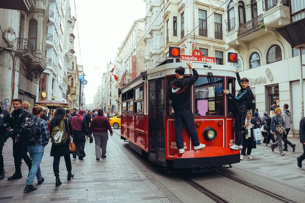 İstiklal Caddesi'nde kırmızı tramvay; arka basamağına tutunmuş gençler, iki yanda kalabalık ve beyaz cepheli eski apartmanlar.
