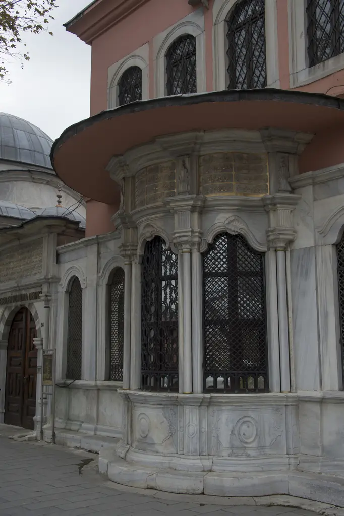 Şah Sultan Sebili; oval mermer gövde, şebekeli üç pencere, üstte çıkıntılı saçak ve pembe mektep cephesi.