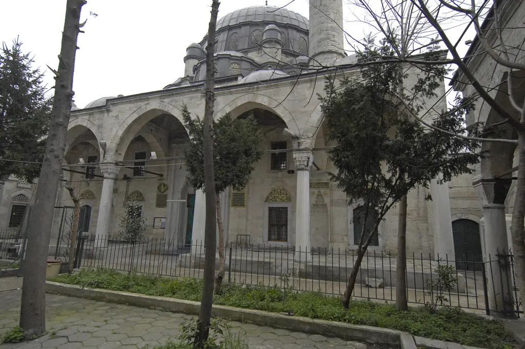 Avludan Nişancı Mehmed Paşa Camii; sütunlu son cemaat revakı, yarım kubbeler, ana kubbe, minare ve önde çıplak ağaçlarla demir parmaklık.