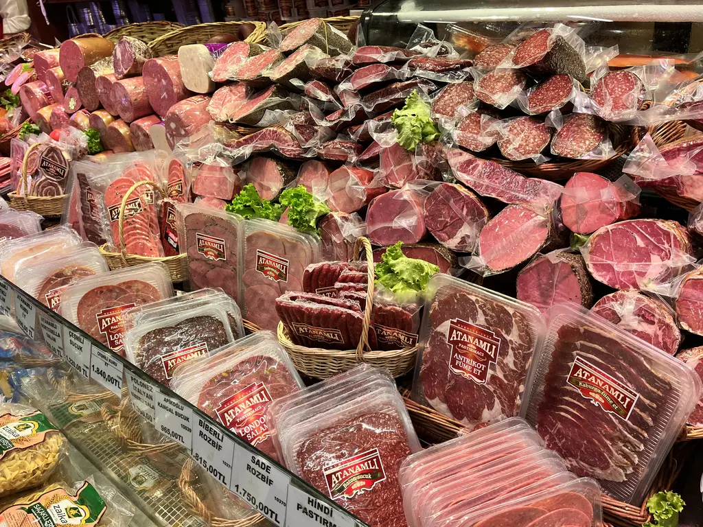 Eminönü'nde bir şarküterinin vitrini; dilimlenmiş pastırma ve sucuklar, paketlenmiş etler ve marul yaprakları.