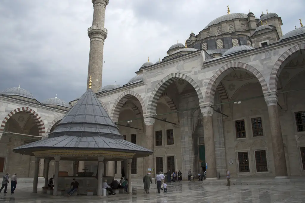 Fatih Camii avlusunda şadırvan yakın planda; koyu gri konik kurşun çatı ile altın alem, arkada kırmızı beyaz almaşık taşlı yüksek revak kemerleri ve bir minare.