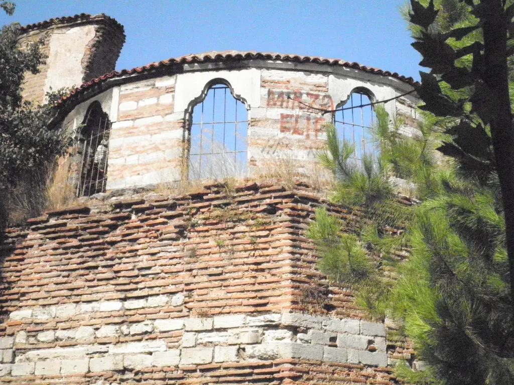 İmrahor İlyas Bey Camii'nin apsisi; dönüşümlü taş ve tuğla sıralı yarım yuvarlak gövde, üstte demir şebekeli üç kemerli pencere, kiremitli saçak ve mavi gökyüzü.