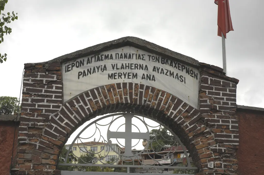 Tuğla örgülü kemerli bahçe kapısı; üstteki beyaz levhada Rumca ve Türkçe "Panayia Vlaherna Ayazması Meryem Ana" yazısı, kemerde demir haç.