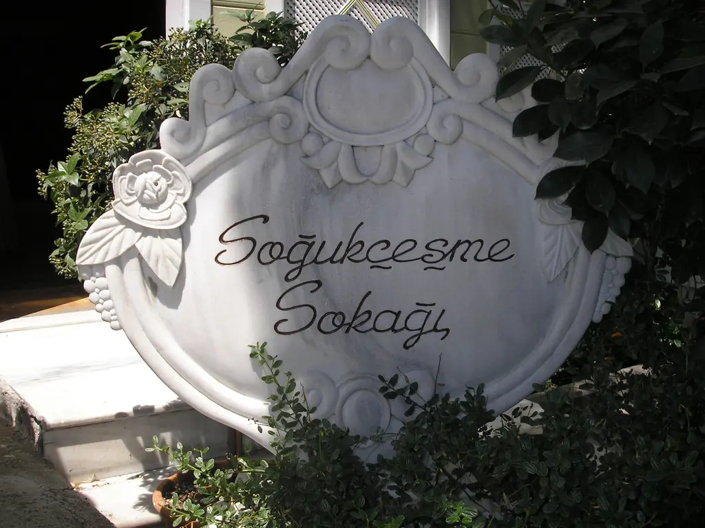 Soğukçeşme Sokağı'nın adı el yazısıyla kazınmış kıvrımlı mermer levhası; çevresinde yeşil yapraklar ve arkada beyaz bir duvar.